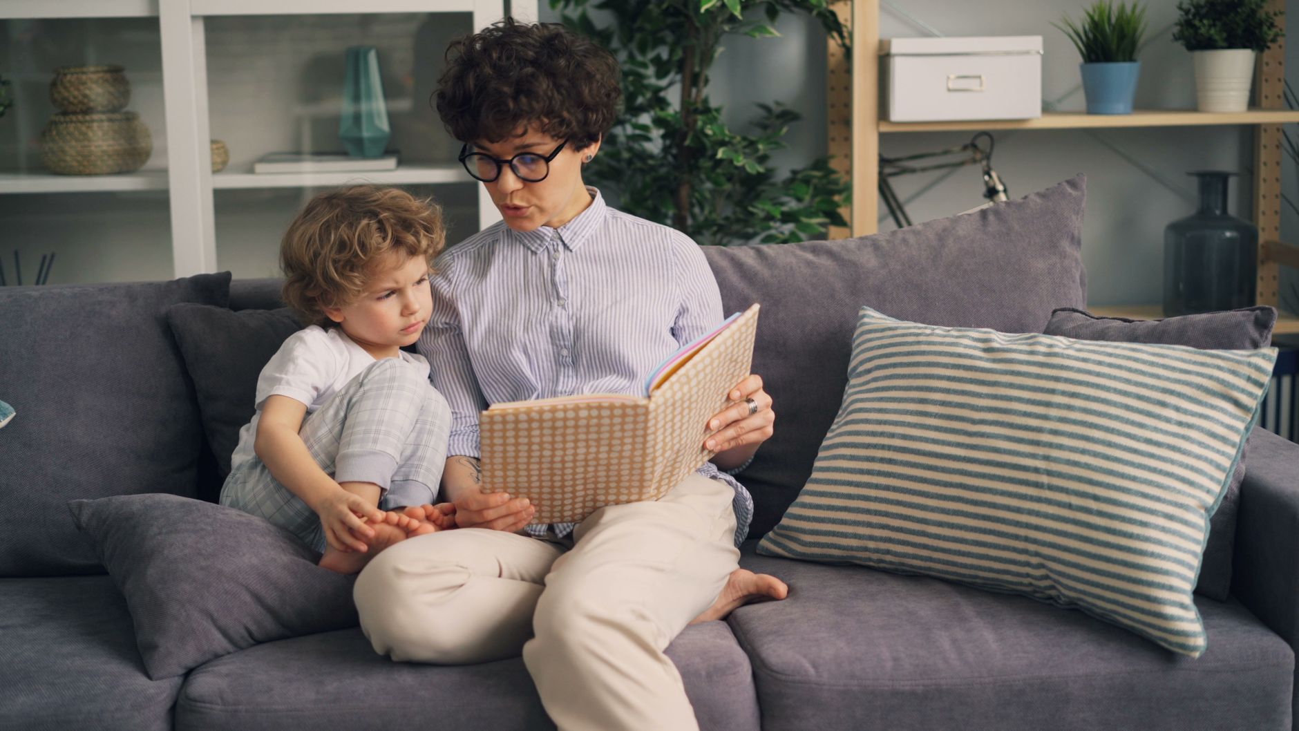 An adult and young child reading a book together on a grey couch.